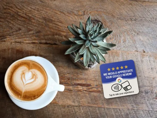 Tarjeta NFC de reseñas de Google apoyada sobre una mesa de madera junto a un café con leche